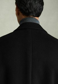 Sort uldfrakke med en struktureret krave, vist fra ryggen, parret med en grå højhalset trøje. Glat tekstur, minimal detaljeringsgrad, moderne design.