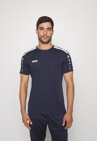 T-shirt sportivo navy a maniche corte, con logo JAKO, strisce bianche sulle spalle e collo rotondo. Presenta punti grigi sulle spalle.