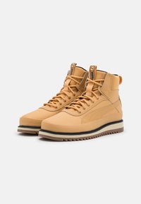 Vysoké tenisky vyrobené z hnědé kůže s texturovaným povrchem, s černou a béžovou podrážkou, kulatou špičkou a výraznými tkaničkami s logovými detaily.