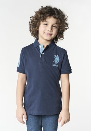 Ragazzo giovane con capelli ricci che indossa una polo blu navy con un logo di cavallo e cavaliere blu e jeans blu, sorridente verso la fotocamera.