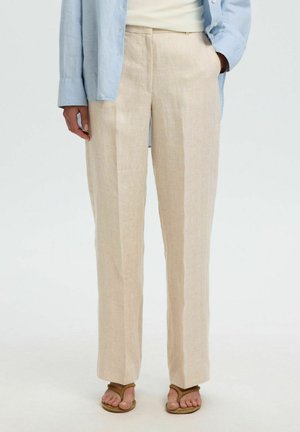 Personne portant un pantalon en lin beige, une chemise bleu clair et des sandales marron, debout avec une main dans la poche devant un fond uni.