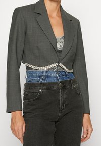 Blazer gris corto con solapas afiladas sobre una blusa de lentejuelas y pantalones de mezclilla en capas en azul y negro, con un detalle de cinturilla joya.