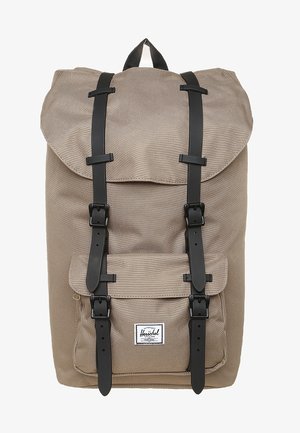 Sac à dos en tissu beige avec des sangles noires à boucle, poche avant et poignée supérieure, avec une étiquette de marque blanche rectangulaire sur la poche.