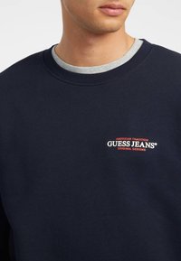 Sudadera de algodón marina con cuello redondo, que presenta un interior de color gris y un logo "GUESS JEANS" en rojo y blanco en el pecho.