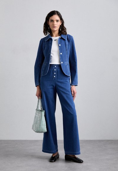 Jeansowa kurtka z ostrym kołnierzem, białymi guzikami i niebieskimi dżinsami o szerokich nogawkach. Trzyma w ręku torbę w zielono-białe paseczki, w połączeniu z czarnymi butami.