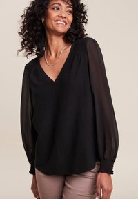 Blouse noire texturée avec un décolleté en V et des manches longues transparentes, dotée de poignets élastiques et d'une coupe floue, associée à un pantalon clair.