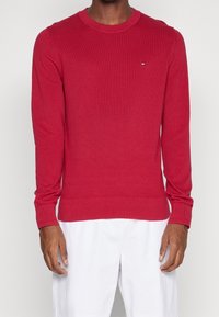 Suéter de punto acanalado rojo con escote redondo, mangas largas ajustadas y un pequeño logotipo en el pecho. Combinado con pantalones blancos.