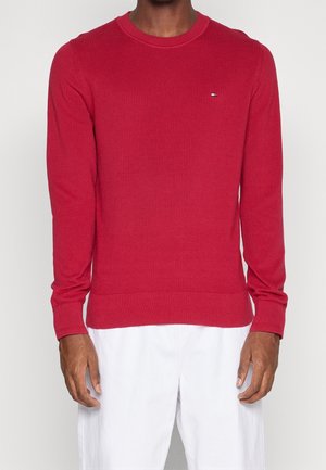 Maglione rosso a coste con scollo rotondo, maniche lunghe aderenti e un piccolo logo sul petto. Abbinato a pantaloni bianchi.