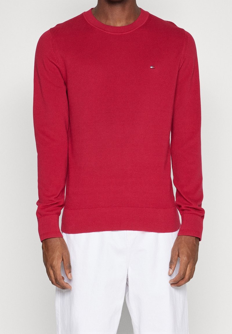 Suéter de punto acanalado rojo con escote redondo, mangas largas ajustadas y un pequeño logotipo en el pecho. Combinado con pantalones blancos.