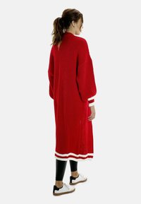 Cárdigan largo de punto rojo con textura acanalada, acentos de rayas blancas en las mangas y el dobladillo, usado sobre leggings negros y zapatos blancos.