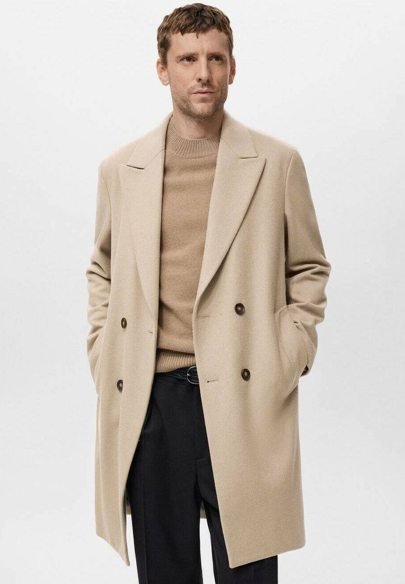 Abrigo de mezcla de lana en beige, de doble botonadura, con grandes solapas, bolsillos laterales y botones oscuros, llevado sobre un suéter de punto beige y pantalones negros.