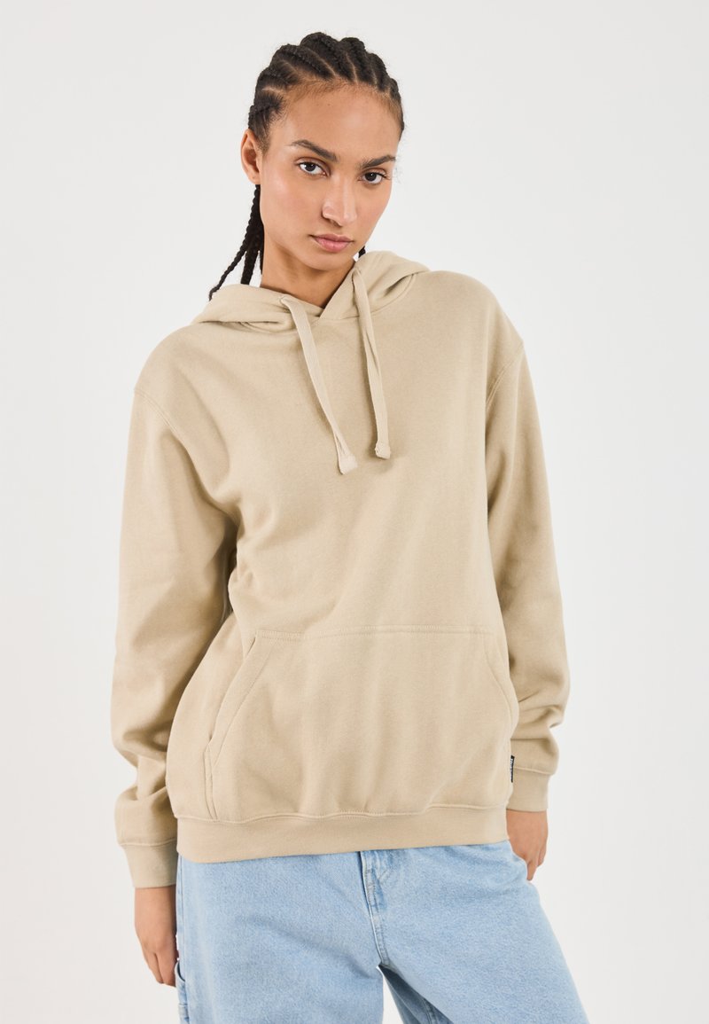 Kvinde med fletninger iført en beige hættetrøje og lyseblå jeans, der står med et neutralt udtryk foran en plain hvid baggrund.