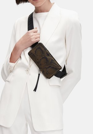 Femme en costume blanc portant un sac bandoulière à motif marron foncé avec une sangle noire réglable croisée sur la poitrine.