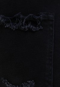 Detailní záběr černé džínoviny se dvěma opotřebovanými, třepenými horizontálními trhlinami blízko okraje přišitých kapes.