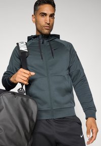 Mørkegrønn zip-up hettegenser med hette, glidelåslommer på sidene og svarte detaljer. Modellen holder en svart sportstrekker med en hvit stropp.