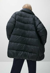 Person med kort blondt hår iført en overdimensioneret sort dunjakke og mørke bukser, stående mod en ensfarvet lys baggrund, med ryggen til kameraet.