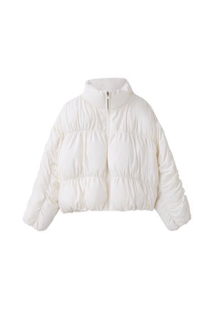 Femme aux cheveux bruns raides porte une doudoune blanche sur un crop top rouge et un pantalon large rouge, debout devant un fond gris et rouge.