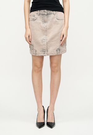 Femeie purtând o mini fustă din denim bej deschis cu buzunare frontale și un top negru fără mâneci, asortate cu pantofi cu toc înalt, negri și ascuțiți.