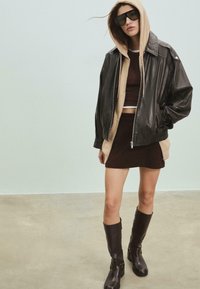 Žena v nadměrném černém koženém saku přes béžovou mikinu, hnědým cropped topem, hnědými sukněmi, vysokých kozačkách a velkých slunečních brýlích v prostorné místnosti.