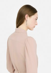 Orecchini pendenti in argento con design geometrico e accenti trasparenti, indossati con una blusa rosa chiaro con colletto e maniche a sbuffo.