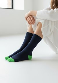 Osoba sedící na podlaze, obutá v tmavě modrých ponožkách se světlounce zelenými špičkami a patami, oblečená do bílého oděvu u okna.