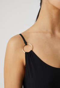 Μαύρο ένδυμα με λεπτό λουράκι που διαθέτει μεταλλικό δαχτυλίδι σε χρυσό χρώμα. Το ύφασμα φαίνεται λείο με ελαφρά κρέμαση κοντά στον ώμο.