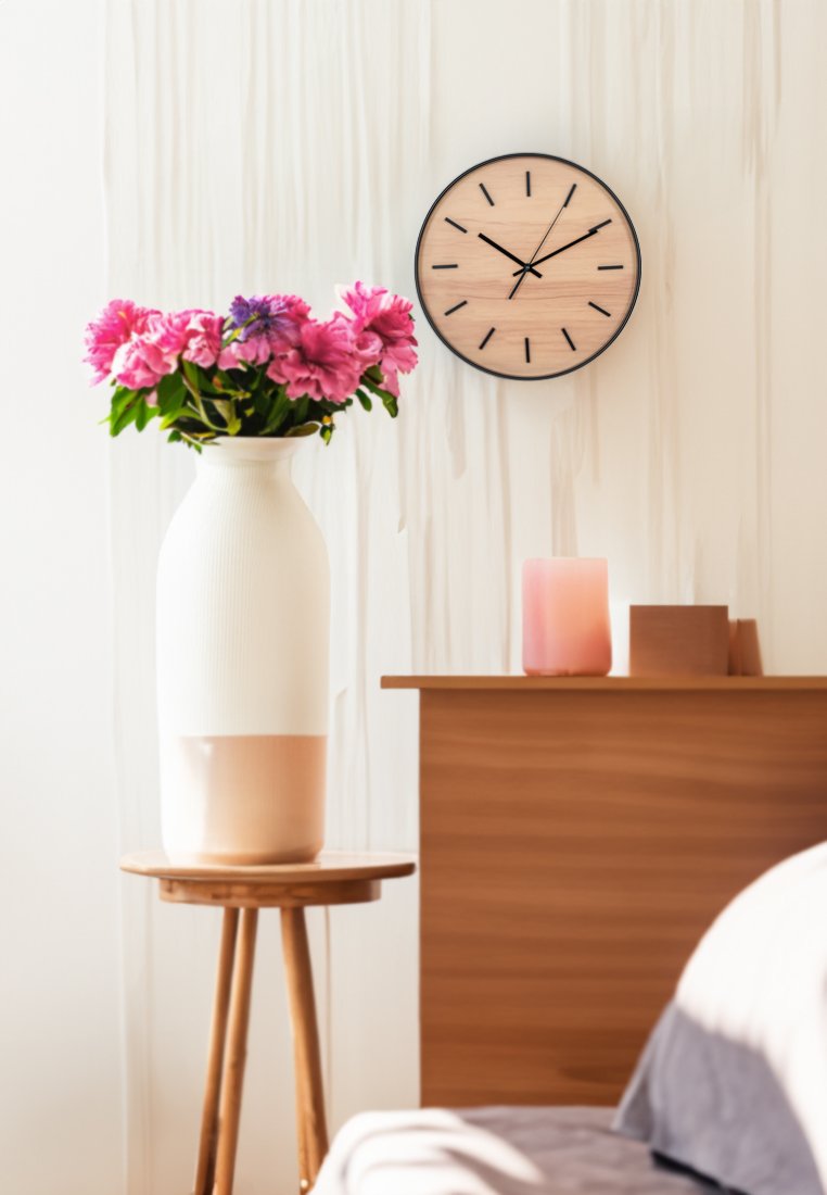 Orologio da parete rotondo con una faccia in legno chiaro, marcature nere e lancette. Nelle vicinanze, un vaso bianco testurizzato con fiori rosa e un comodino in legno.
