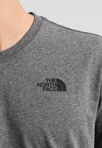 Camiseta de algodón gris con cuello redondo y mangas cortas, que presenta un logotipo negro de "The North Face" en el lado izquierdo del pecho.