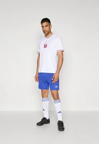 Hvit fotballdrakt med røde og blå detaljer, blå shorts, hvite knelange sokker med blå striper, og svarte fotballsko.