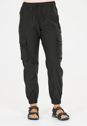 Sorte cargobukser med elastisk talje og manchetter, flere lynlåse og lommer med løg, båret med sorte sandaler med åben tå på en lys baggrund.