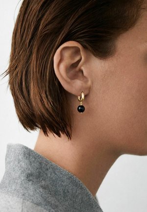 Közelkép egy személy füléről, akinek rövid barna haja van, arany karika fülbevalót visel fekete gyönggyel, és szürke gyapjú gallér látható.