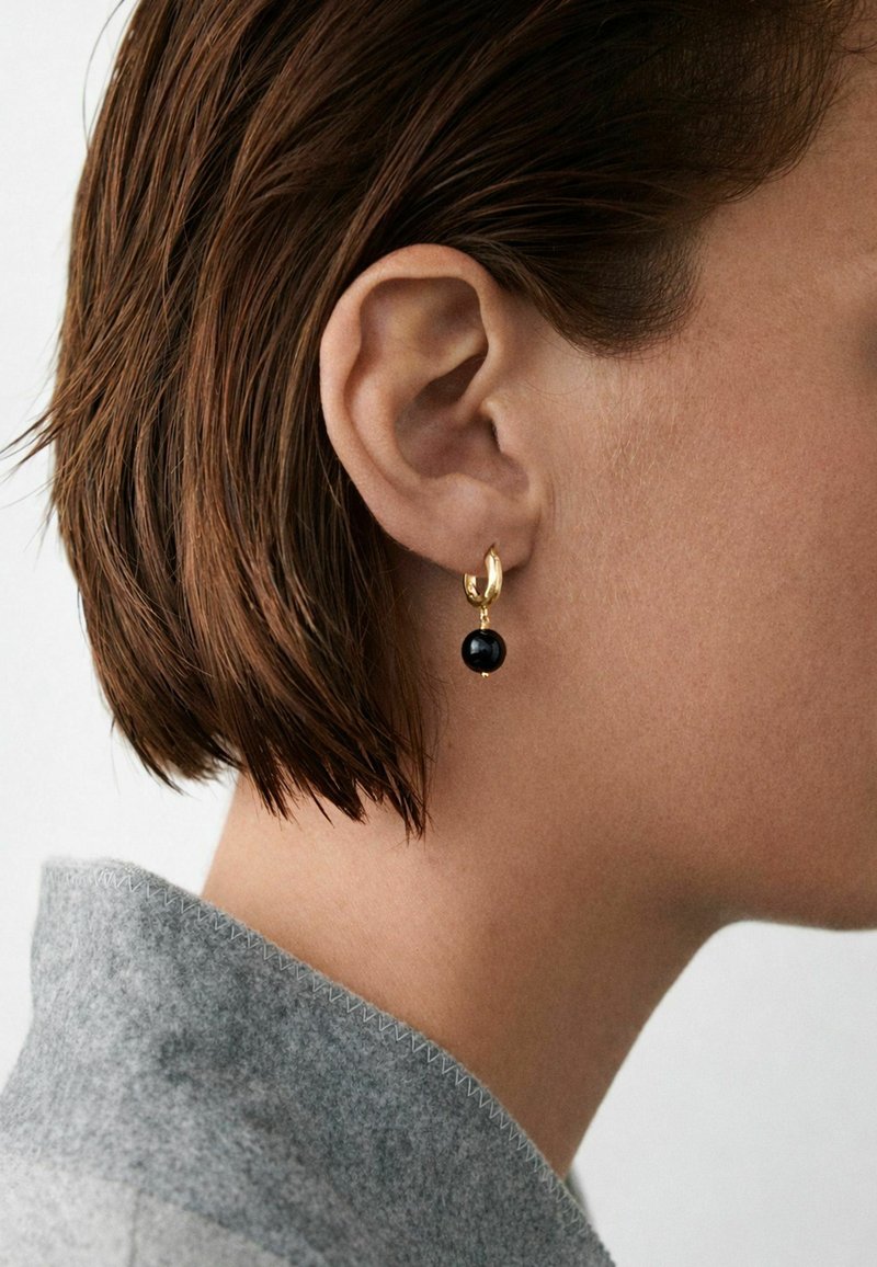 Približni prikaz uha osobe sa kratkom smeđom kosom koja nosi zlatnu naušnicu na obruč s crnom perlicom i vidljivim sivim vunenim ovratnikom.