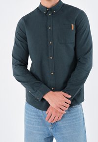 Uomo che indossa una camicia verde scuro con bottoni e taschino sul petto e jeans azzurri chiari, con le mani incrociate davanti, su sfondo bianco.