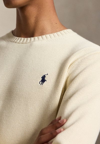 Lähivõte inimesest, kes kannab kreemjat kootud kampsunit tumesinise tikitud polomängija logoga rinnal.