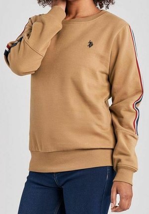 Personne portant un sweat-shirt beige avec des bandes rouges, blanches et noires sur les manches et un petit logo noir sur la poitrine, associé à un jean bleu foncé.