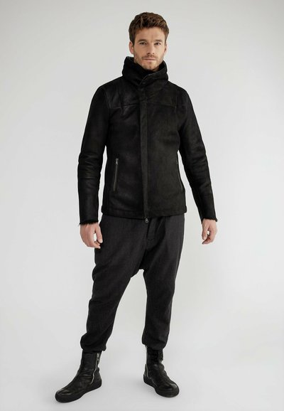 Schwarze Wildlederjacke mit hohem Kragen, mit Reißverschluss versehenen Taschen und strukturierter Oberfläche, kombiniert mit dunkel gefärbten, locker sitzenden Hosen und schwarzen Stiefeln.