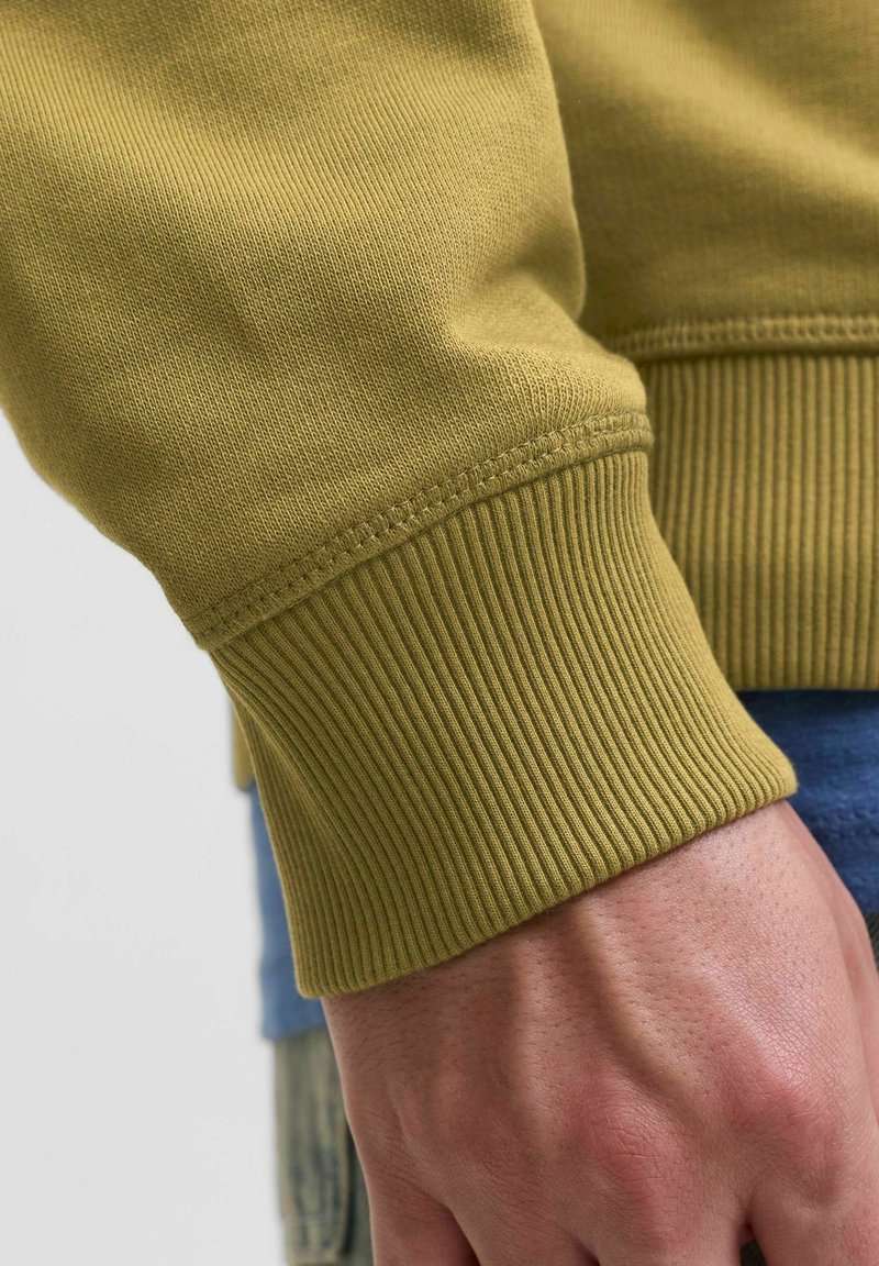 Poignet de sweat-shirt vert olive avec texture en côtes, présentant un ourlet cousu et une surface lisse. Main partiellement visible à côté.