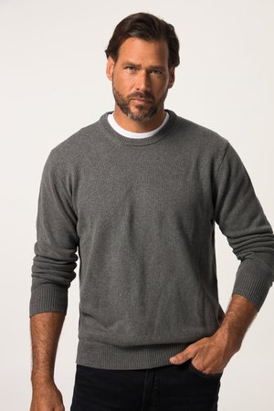 Mand med skæg iført en grå langærmet sweater og sorte bukser, stående med den ene hånd i lommen mod en ensfarvet baggrund.