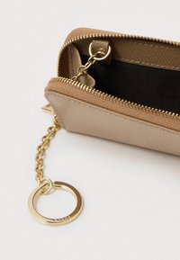 Tan kunstlæderpung med gyldent lynlås, kæde og nøglekrog. Indvendig har den en sort foring. Kompakt design med tekstureret overflade.