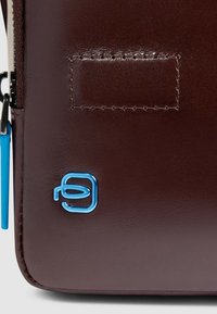 Braune Ledertasche mit glatter Textur, blauem Logoakzent und sichtbarer Nahtdetail in der Nähe des Reißverschlusses.