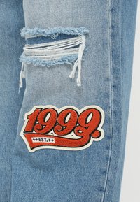 Világoskék farmernadrág, amelynek térdén kopott rész található, széttépett szélekkel, és egy hímzett, piros és fehér „1999” feliratú folt.