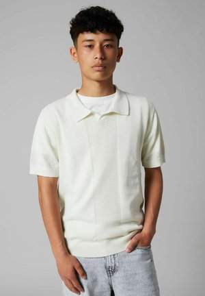 Nuori mies, jolla on lyhyet kiharat hiukset, pukeutuneena valkoiseen teksturoituun lyhythihaiseen pooloneuleeseen ja vaaleanharmaisiin farkkuihin, seisoo käsi yhdessä taskussa.