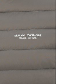 Geacă gri căptușită cu secțiuni orizontale quilted. Prezintă un logo alb care scrie "ARMANI EXCHANGE MILANO / NEW YORK" în față.