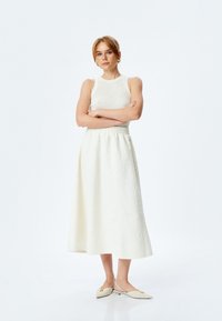 Jupe midi blanche texturée avec une taille ajustée, associée à un top sans manches fitted en tricot, et des chaussures slip-on beiges avec des accents dorés.