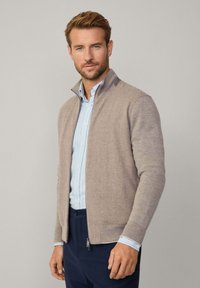 Pull zippé marron clair avec un design de tricot texturé, un col montant et des couleurs de manches contrastantes, associé à une chemise rayée bleu clair.
