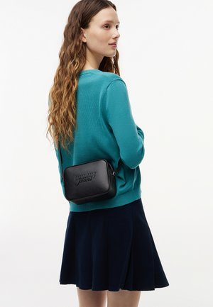Donna con capelli lunghi e mossi indossa un maglione turchese, una gonna blu scuro e una borsa a tracolla nera Tommy Jeans su uno sfondo chiaro e semplice.