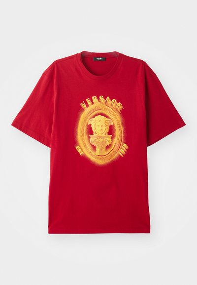 Raudona trumpa rankovė T-marškiniai su geltona Medūzos galvos emblema, apsupta ovalo, su užrašu "VERSACE" ir "EST. 1978" priekyje.