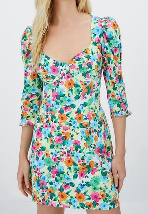 Vrouw draagt een aansluitende bloemenjurk met pofmouwen tot driekwart lengte en een sweetheart halslijn in felle roze, oranje, blauwe en groene kleuren.