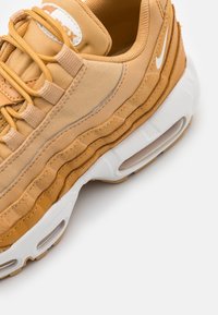 Gros plan sur une chaussure de sport beige et blanche avec des textures superposées, une semelle épaisse et un coussin d'air visible dans la semelle extérieure.