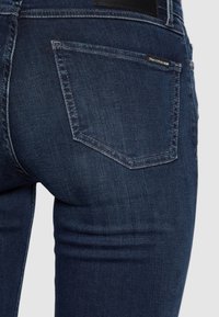 Jeans in denim blu scuro con una vestibilità slim, caratterizzati da una singola tasca posteriore e dettagli di cucitura discreti. Etichetta in pelle visibile sulla cintura.